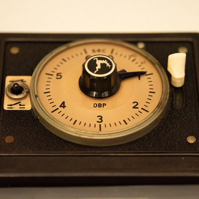 The image features a small, old-fashioned analog clock with a black and gold face. It is placed on top of a table or counter, possibly in a room with white walls. The clock has a dial that reads 14 degrees DP (degrees per minute) and a needle pointer at the center.