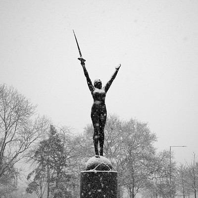 The image features a statue of a woman holding a sword, standing on top of a pedestal. She is positioned in the center of the scene and appears to be looking upwards. The statue is surrounded by trees, with some located near the base of the pedestal and others further away.