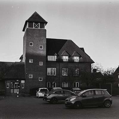 The image is a black and white photo of a parking lot in front of a large brick building. There are several cars parked in the lot, with some closer to the foreground and others further back. A fire station can be seen nearby, adding an interesting element to the scene.
