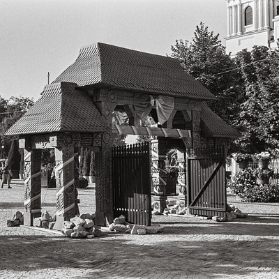The image is a black and white photo of an outdoor structure, possibly a gazebo or a small building. It has a thatched roof and is surrounded by trees. There are several people in the scene, some standing near the structure while others are further away.