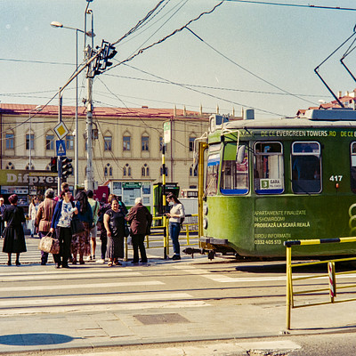 The image depicts a busy city street with a green trolley car driving down the road. There are several people standing on the sidewalk, waiting to board or just observing the scene. Some of them have handbags and backpacks, indicating they might be commuters or travelers.