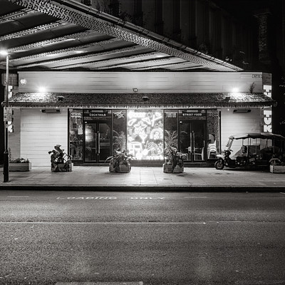 The image is a black and white photo of a street scene at night. There are several motorcycles parked on the sidewalk, with one near the center of the frame and two others closer to the left edge. A car can be seen in the background, parked further down the road.