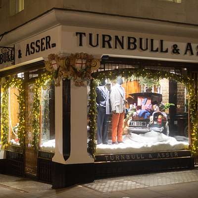 The image features a storefront of a clothing store called Turnbull & Asser. The store has a large window display that showcases various mannequins wearing different outfits, creating an eye-catching visual for passersby. There are also several potted plants placed around the storefront, adding to the overall ambiance of the shop.