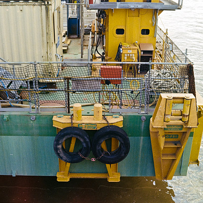 The image features a large yellow boat with a green top, docked in the water. On the front of the boat, there are two black tires and a metal barrier. Additionally, there is a ladder on the side of the boat for easy access to the deck.