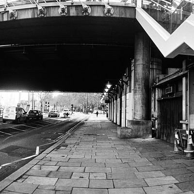 The image is a black and white photo of an underpass with a bridge above it. There are several cars driving on the road, including one truck. A few people can be seen walking along the sidewalk near the bridge.