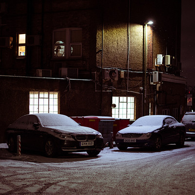 The image depicts a snowy night scene with several cars parked on the side of a street. There are three cars in total, one being closer to the left edge of the frame and two others positioned more towards the center. A truck is also visible near the right edge of the image.