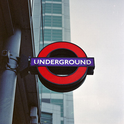The image features a large sign for the London Underground, which is prominently displayed on a building. The sign has a red background with white letters that spell out "Underground." In addition to the main sign, there are several smaller signs visible in the scene, likely providing additional information or directions related to public transportation.