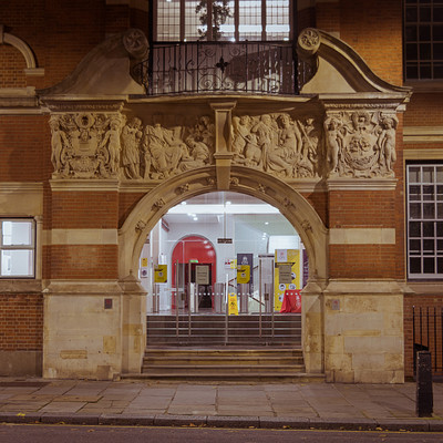 The image features a large, red brick building with an arched entrance. Above the entrance, there are several statues and carvings adorning the facade of the building. The street outside is lined with benches for people to sit on while enjoying their surroundings.