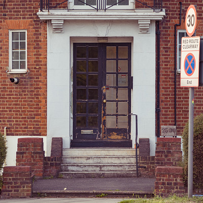 The image features a red brick building with a black door, which is the main focus of the scene. Above the door, there are two signs: one is a "Red Route Clearway" sign and the other is a "No Right Turn" sign. In front of the building, there are steps leading up to the entrance.