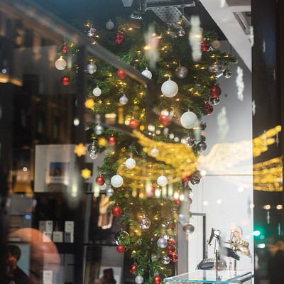 The image features a store window with a beautifully decorated Christmas tree in the center. The tree is adorned with ornaments, including red and white balls, creating an eye-catching display. There are also several people visible outside the store, likely admiring the festive scene or passing by.