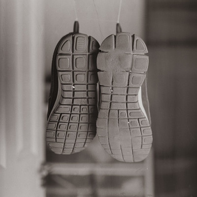 The image is a black and white photo of two pairs of shoes hanging on the wall. One pair of shoes is located towards the left side, while the other pair is positioned more towards the center-right area of the frame. The shoes are suspended from hooks or pegs, creating an interesting visual effect.