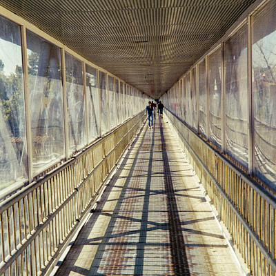 The image is a black and white photo of an empty pedestrian bridge. There are two people walking on the bridge, one closer to the left side and another further back on the right side. The bridge has a metal railing along its length, providing safety for those using it.