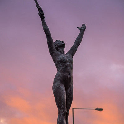 The image features a statue of a woman holding her arms up in the air, possibly celebrating or expressing joy. She is standing on top of a pole, which has a street light attached to it. The scene takes place during sunset, with warm colors illuminating the sky and casting a soft glow over the statue.