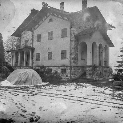 The image is a black and white photo of an old house with a large chimney. It appears to be a winter scene, as the house has snow on it. There are several steps leading up to the front door of the house.