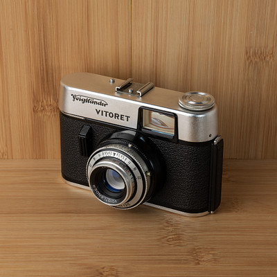 The image features a vintage camera, likely an old Kodak Vitoret model, sitting on top of a wooden table. The camera is black and silver in color, with its lens facing the viewer. It appears to be an antique or classic piece, possibly used for photography in the past.