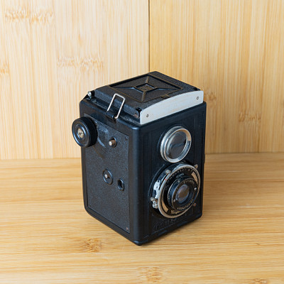 The image features a black and white vintage camera sitting on top of a wooden table. The camera is an old-fashioned model, possibly a Polaroid or a similar type of instant camera. It has a strap attached to it, which could be used for carrying the camera around.