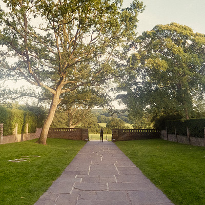The image features a beautiful garden with a tree in the center, surrounded by a brick path. The path is lined with trees and bushes on both sides, creating an inviting atmosphere for visitors to enjoy the outdoors. There are several benches placed along the path, providing ample seating options for people to relax and take in the scenery.