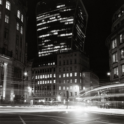 The image is a black and white photo of a city street at night, featuring a tall building with many windows. There are several cars driving down the street, creating a sense of motion in the scene. Traffic lights can be seen along the road, indicating that this is an urban environment with controlled intersections.