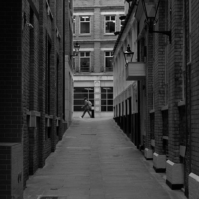 The image is a black and white photo of an alleyway between two buildings. A man can be seen walking down the alley, while another person stands on the sidewalk near one of the buildings. There are several windows visible in the scene, with some located above the people and others along the sides of the buildings.