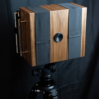 The image features a wooden box with a black background. Inside the box, there is a camera mounted on a tripod, ready for use. The camera appears to be an old-fashioned model, adding a vintage touch to the scene.