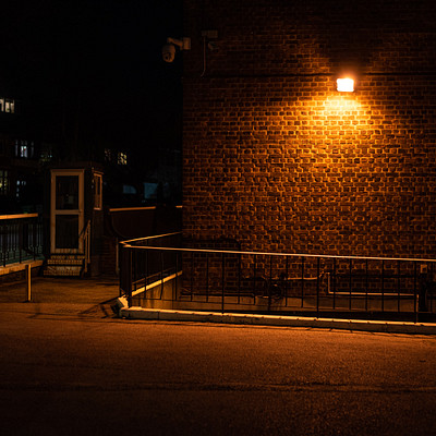 The image is a black and white photo of an empty street at night. A brick building with a light on in the corner serves as the main focus of the scene. There are two benches located near the building, one closer to the left side and another further back on the right side.