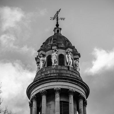 The image features a large, ornate clock tower with a weather vane on top. The clock is located towards the center of the tower and appears to be quite prominent in size. There are four statues adorning the sides of the tower, each one representing an angelic figure.