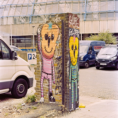The image features a street scene with two colorful graffiti-covered brick pillars, each adorned with a unique design. One of the pillars has a face painted on it, while the other one is decorated with a smiley face. In addition to these eye-catching artworks, there are several cars parked along the street, including two vans and three trucks.