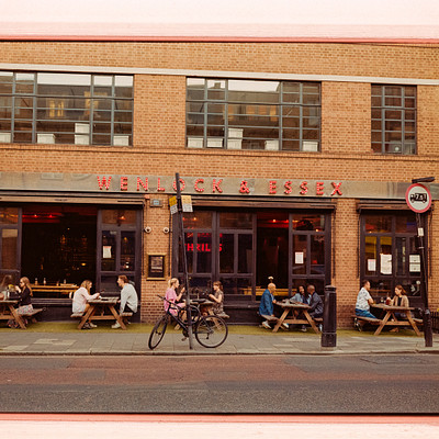 The image is a black and white photo of a street scene with several people sitting at tables outside of a restaurant. There are multiple benches placed around the area, providing seating for the patrons. A bicycle can be seen parked near one of the benches.