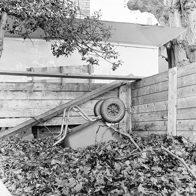 The image features a wheelbarrow with its back tire turned upside down, sitting in the middle of a pile of leaves. The wheelbarrow is positioned on top of a wooden fence or wall, creating an interesting visual effect. There are also some benches visible in the scene, one located near the left side and another further to the right.