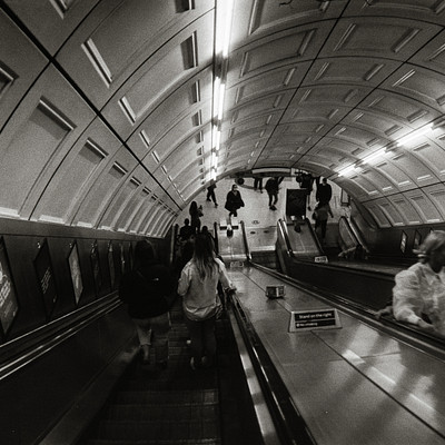 The image is a black and white photo of an underground subway station. There are several people walking around the platform, some carrying handbags or backpacks. A total of 13 people can be seen in various positions throughout the scene.