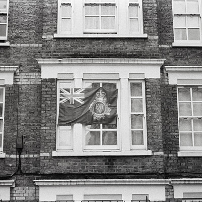 The image is a black and white photo of an old building with a flag hanging from the window. The flag has a red cross on it, which suggests that it might be related to the British military or government. The building appears to have a brick facade, giving it a classic and historical appearance.