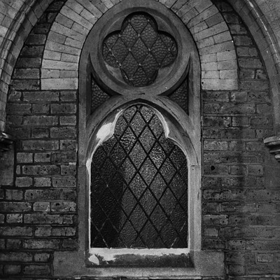 The image is a black and white photo of an old church with a large stained glass window. The window features a cross design, adding to the historical atmosphere of the building. The stone wall surrounding the window adds to its prominence in the scene.