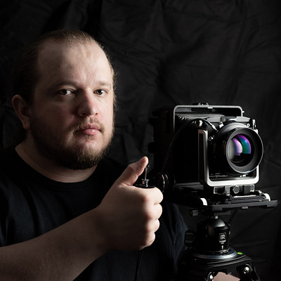 The image features a man with a beard and mustache, wearing a black shirt. He is holding a camera in his hand, giving the impression that he might be taking a picture or preparing to do so. The man appears to be smiling as he holds up the camera, possibly enjoying the moment or capturing an interesting subject.