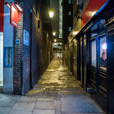 The image depicts a narrow alleyway between two buildings, with one of the buildings being a restaurant. The alley is illuminated by street lamps on both sides, creating a dark atmosphere. A person can be seen standing near the entrance to the restaurant, possibly waiting for someone or preparing to enter.