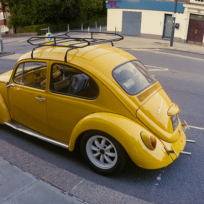 The image features a yellow Volkswagen Beetle parked on the side of a street. It is positioned near the curb, and there are several people in the scene. One person can be seen standing close to the car, while two others are further away from it.