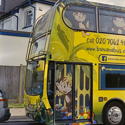 The image features a yellow and blue bus parked on the side of a street. The bus is decorated with monkey faces, giving it a unique appearance. There are several people around the bus, some standing closer to the front while others are further away.