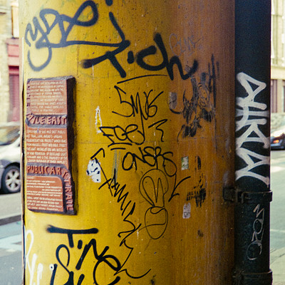 The image features a yellow pole with graffiti covering its surface. There are several cars parked on the street, including one close to the left side of the pole and two others further away on the right side. A parking meter is also visible near the center of the scene.
