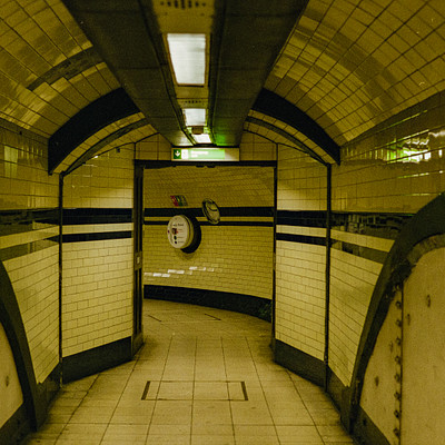 The image is a black and white photo of an underground subway station. There are two clocks visible in the scene, one on the left side and another on the right side of the platform. A person can be seen standing near the center of the platform, possibly waiting for their train or just passing through.