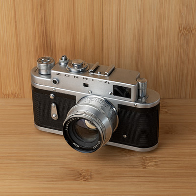 The image features a vintage silver and black camera sitting on top of a wooden table. The camera is an old-fashioned model, possibly a Kodak, with its lens facing the viewer. It appears to be well-maintained and ready for use.