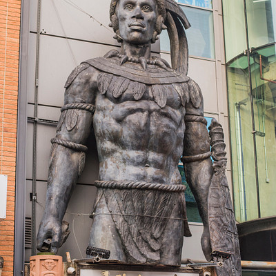 The image features a large statue of a man, possibly an Indian warrior or a similar figure, standing outside in front of a building. The statue is positioned on the side of the building and appears to be made of metal. In addition to the main statue, there are several potted plants placed around the area, adding some greenery to the scene.