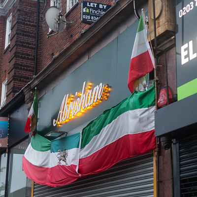 The image features a storefront with a large, colorful flag hanging from the building. The flag is made up of several different colored stripes and has a red, white, and green design. Above the flag, there are two signs on display, one of which reads "Aberdeen." The storefront also displays a few other flags, adding to its vibrant appearance.