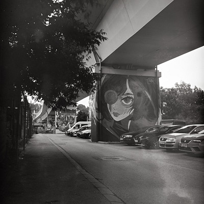 The image is a black and white photo of a street scene with several cars parked on the side. There are at least nine cars visible in various positions along the road, some closer to the foreground while others are further away. A person can be seen walking down the street near one of the cars.