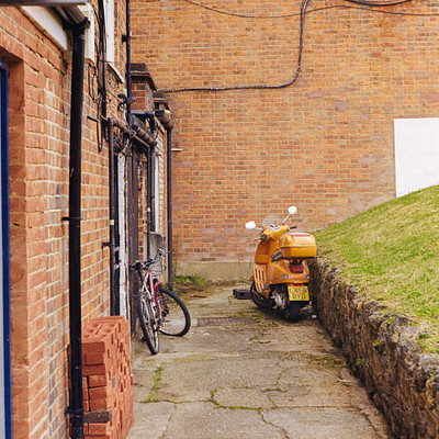 The image features a small brick building with a motorcycle parked in front of it. The motorcycle is positioned on the sidewalk, and there are two bicycles nearby, one closer to the left side of the scene and another further back on the right side. A person can be seen standing near the middle of the scene, possibly observing the surroundings or attending to their vehicle.