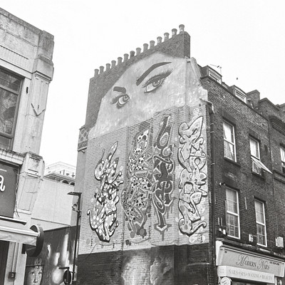 The image is a black and white photo of a building with graffiti on its side. There are several people in the scene, some standing near the building while others are further away. A car can be seen parked nearby, adding to the urban atmosphere of the scene.