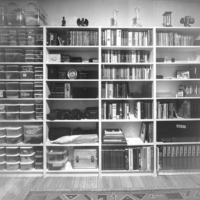 The image is a black and white photo of a bookshelf filled with various books. There are numerous books on the shelves, some placed horizontally while others are stacked vertically. A clock can be seen in the middle of the bookshelf, adding to its decorative elements.