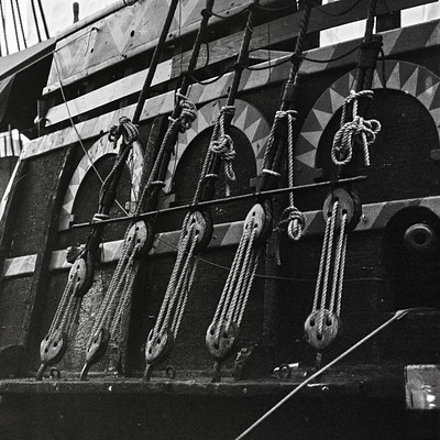 The image is a black and white photo of an old sailboat with its sails down. There are several ropes attached to the boat, likely for securing it or controlling its movement. In total, there are 13 ropes visible in various positions around the boat.