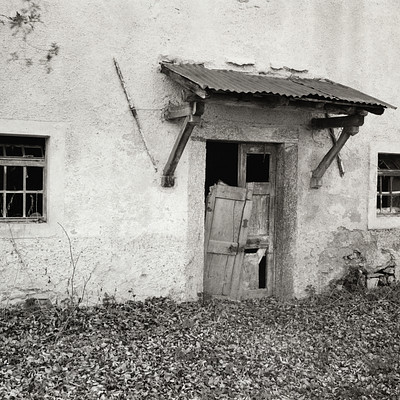 The image is a black and white photo of an old, dilapidated building with a doorway that has been left open. A small shed or shack can be seen attached to the side of the house. There are two windows on the front of the building, one on each side.