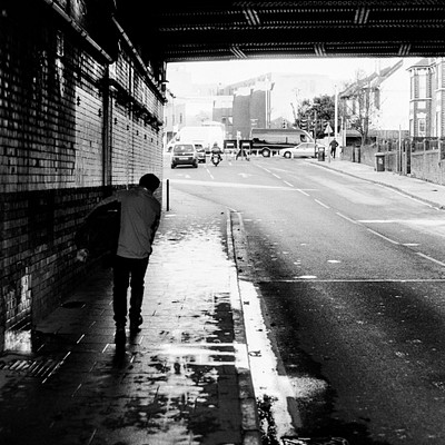 The image is a black and white photo of a man standing on the sidewalk next to a road. He appears to be looking at something in his hand, possibly a map or a piece of paper. There are several cars parked along the street, with one car closer to the foreground and others further down the road.