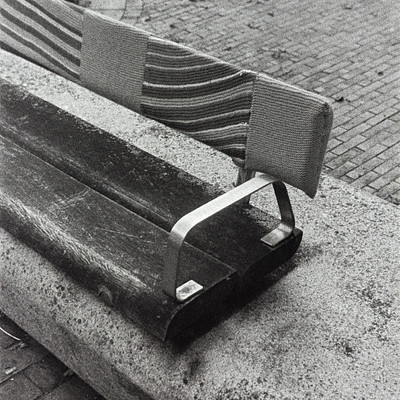 The image is a black and white photo of an empty park bench. It is located on the sidewalk, with a brick road nearby. The bench appears to be made of wood and has a metal armrest.