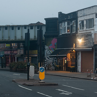 The image depicts a city street at night, with a train crossing over the road. There are several shops and businesses lining the street, including a restaurant on one side of the road. A few cars can be seen parked along the street, and there is a bicycle leaning against a wall near the center of the scene.